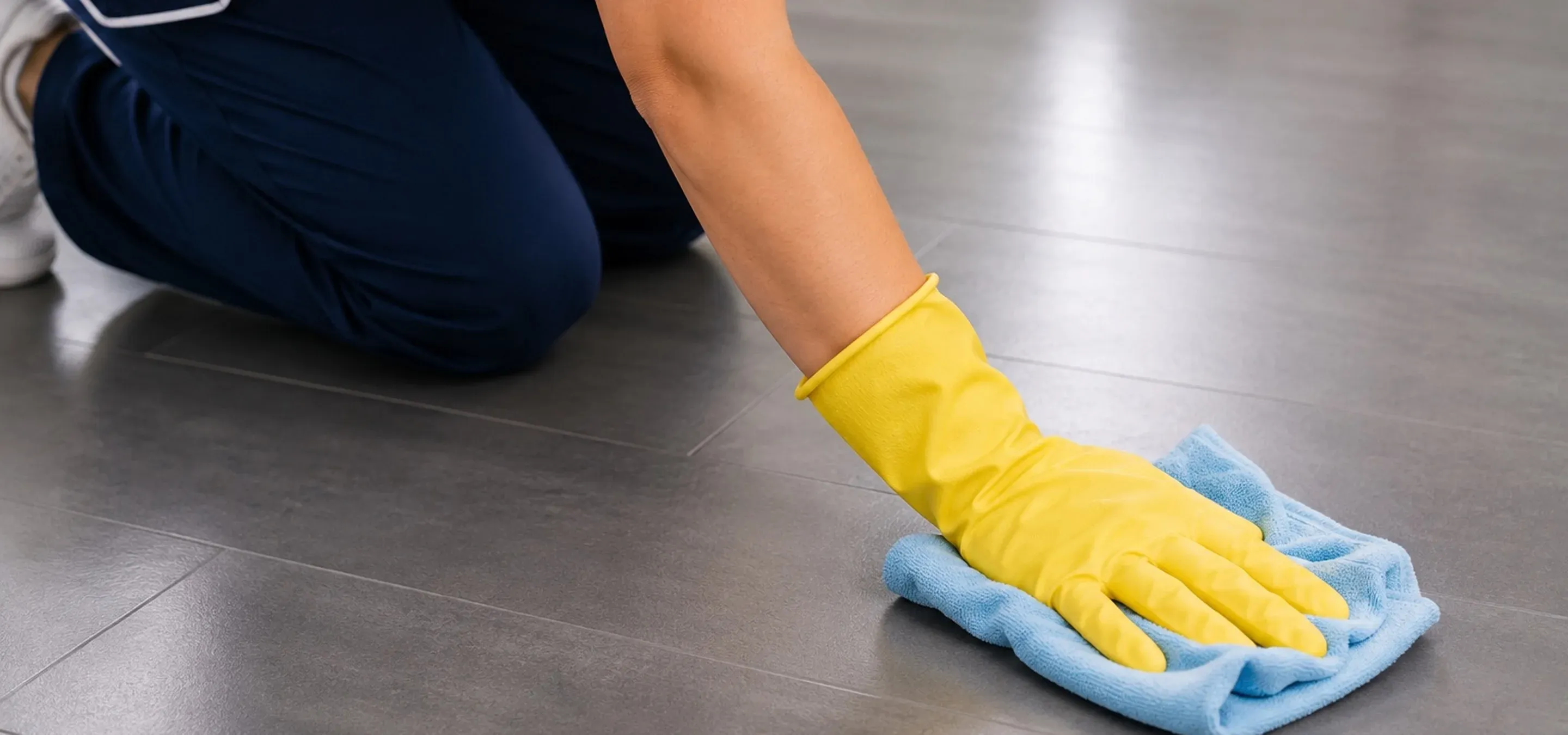 Reinigungskraft wischt einen grauen Fliesenboden mit Mikrofasertuch und gelbem Schutzhandschuh